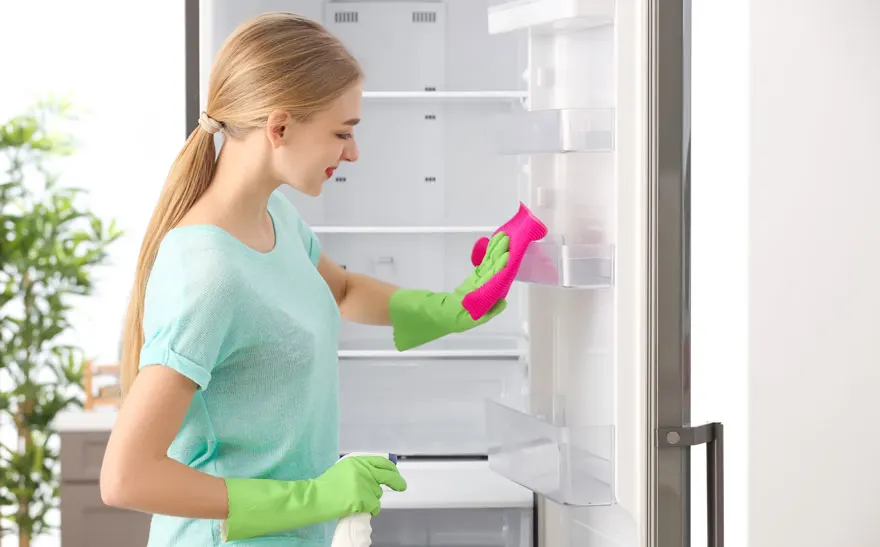 Une personne nettoie un réfrigérateur blanc avec des gants verts et un chiffon rose. Un vaporisateur est visible.