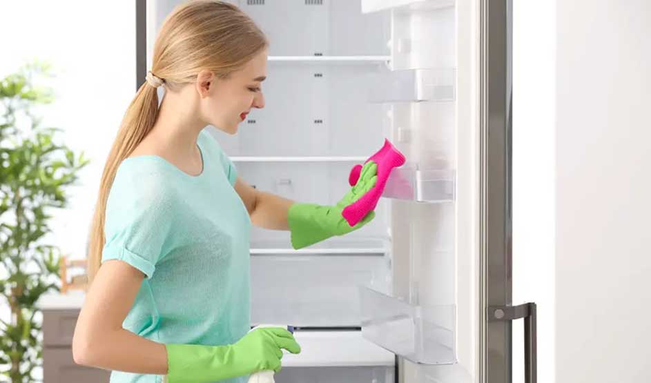 Een koelkast wordt schoongemaakt met een roze doek en groene handschoenen in een keuken.