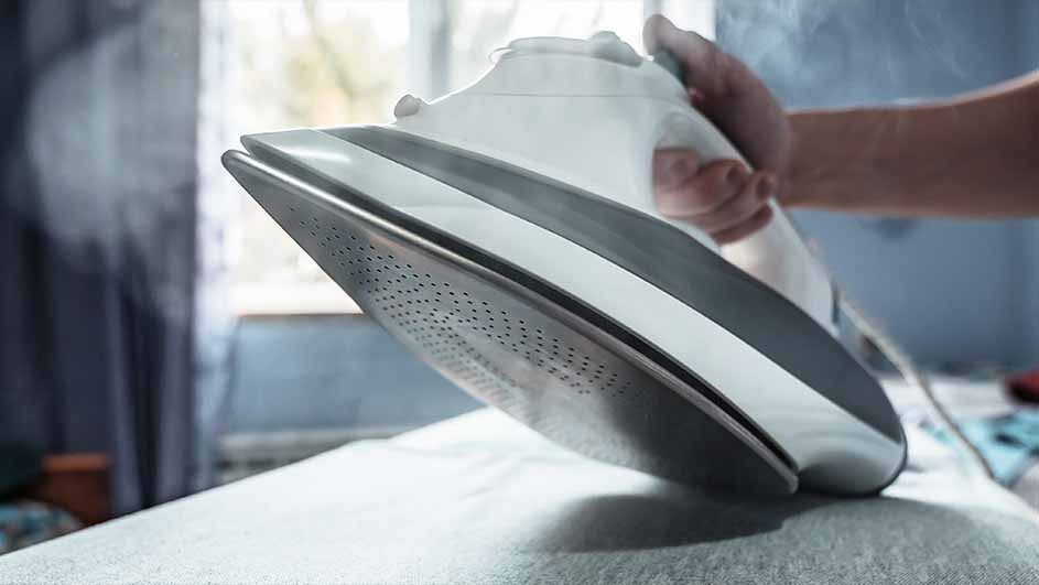 Un fer à repasser blanc et gris, produisant de la vapeur, est posé sur un tissu blanc, avec une fenêtre en arrière-plan.