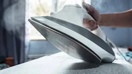 Un fer à repasser blanc et gris, produisant de la vapeur, est posé sur un tissu blanc, avec une fenêtre en arrière-plan.