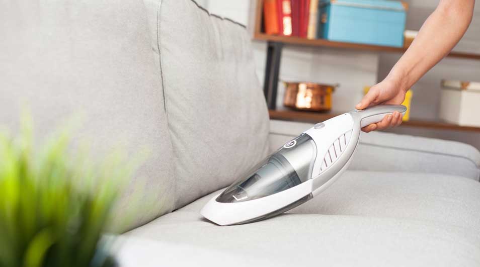 Een grijze sofa wordt schoongemaakt met een witte kruimeldief. Op de achtergrond is een houten rek te zien.