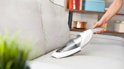 Een grijze sofa wordt schoongemaakt met een witte kruimeldief. Op de achtergrond is een houten rek te zien.