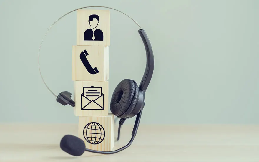 Casque noir près de blocs de bois avec icônes : personne, téléphone, e-mail et globe. Fond uni clair.