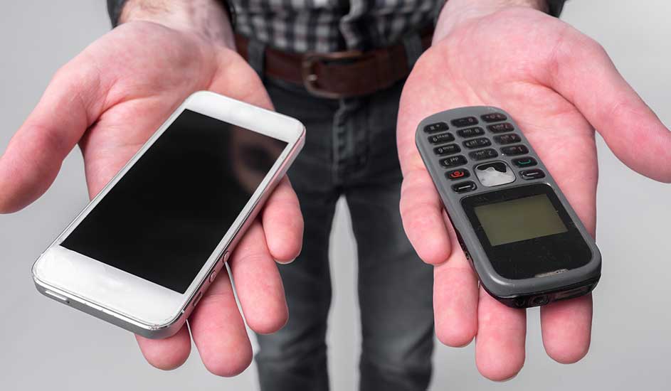 Een persoon houdt een witte smartphone en een grijze feature phone vast, tegen een achtergrond van een geruit hemd en jeans.