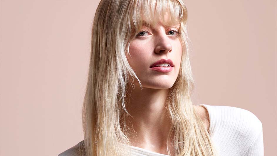 Portrait d'une femme aux longs cheveux blonds et à la frange, portant un haut blanc sur fond beige uni.