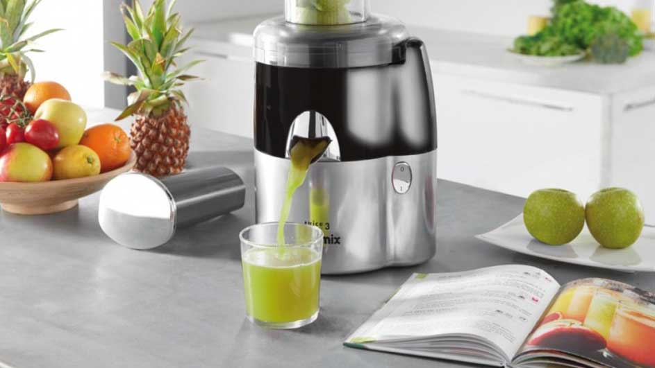 Un presse-agrumes en inox verse du jus vert dans un verre. Un bol de fruits, des pommes et un livre de cuisine sont sur le comptoir.