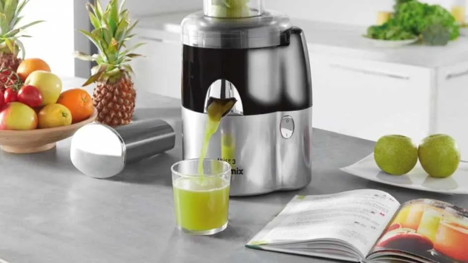 Un presse-agrumes en inox verse du jus vert dans un verre. Un bol de fruits, des pommes et un livre de cuisine sont sur le comptoir.