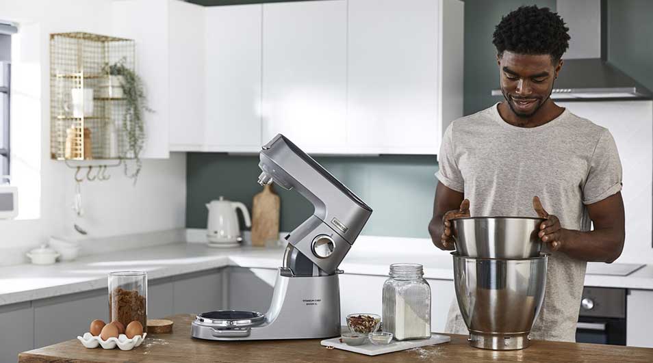 Een zilveren keukenrobot staat op een houten aanrecht in een keuken. Er staan ook eieren, bloem en een kom.