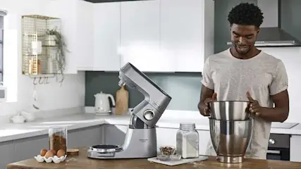 Een zilveren keukenrobot staat op een houten aanrecht in een keuken. Er staan ook eieren, bloem en een kom.