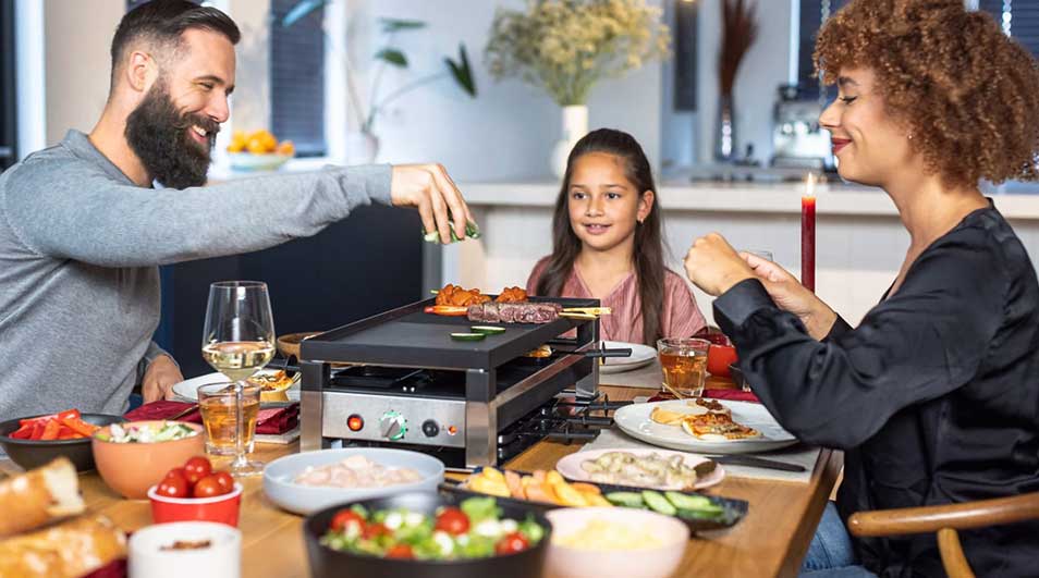 Une table à manger avec un appareil à raclette central, entourée d'aliments variés et de boissons.