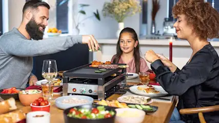 Une table à manger avec un appareil à raclette central, entourée d'aliments variés et de boissons.