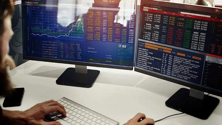 Twee monitoren tonen aandelenmarktgegevens en grafieken. Een toetsenbord en muis staan op een witte tafel.