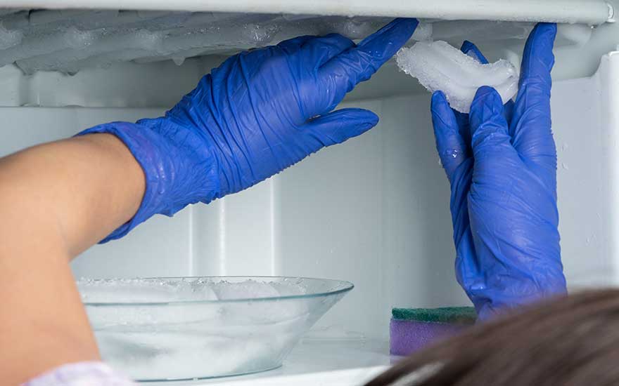 Des mains gantées de bleu retirent de la glace d'un congélateur blanc. Un bol en verre et une éponge sont visibles.