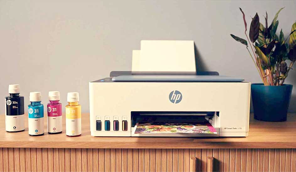 Imprimante HP blanche sur table en bois avec bouteilles d'encre noire, cyan, magenta et jaune. Pot de plante verte à droite.