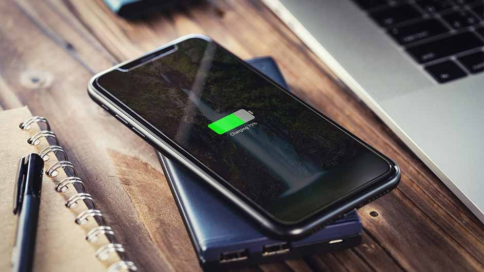 Un téléphone noir en charge sur une batterie externe bleue, à côté d'un ordinateur portable et d'un bloc-notes sur une table en bois.