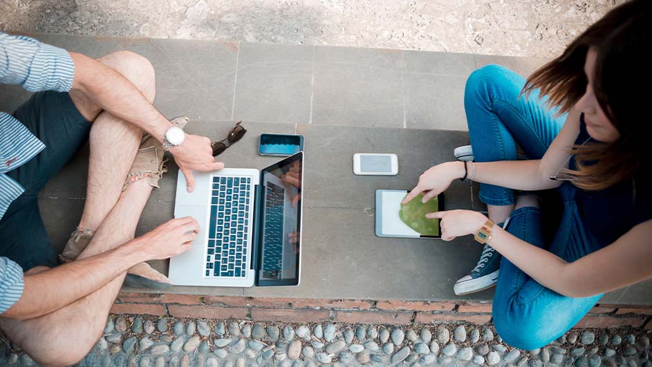 Twee mensen zitten buiten met een laptop, tablet en telefoons op een grijze stenen ondergrond met een kiezelstenen rand.