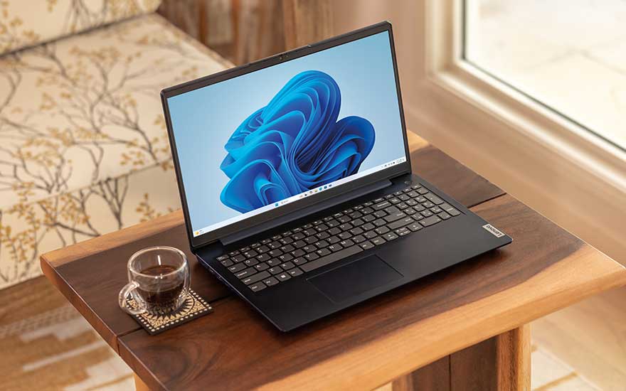 Een zwarte laptop met Windows 11‑scherm staat op een houten bijzettafel naast een glas koffie, bij het raam in een gezellige kamer.