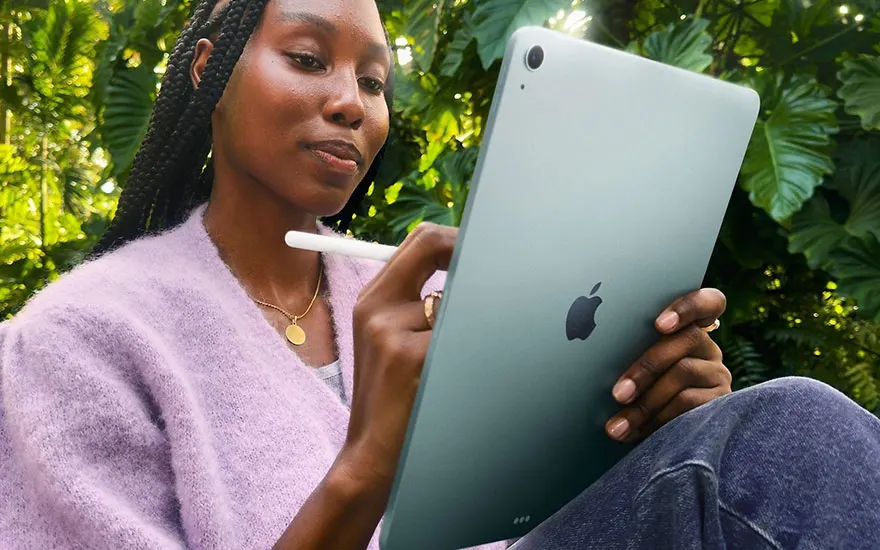 Vrouw in roze trui houdt groene iPad vast terwijl ze buiten zit tussen weelderige groene planten en gefilterd zonlicht.