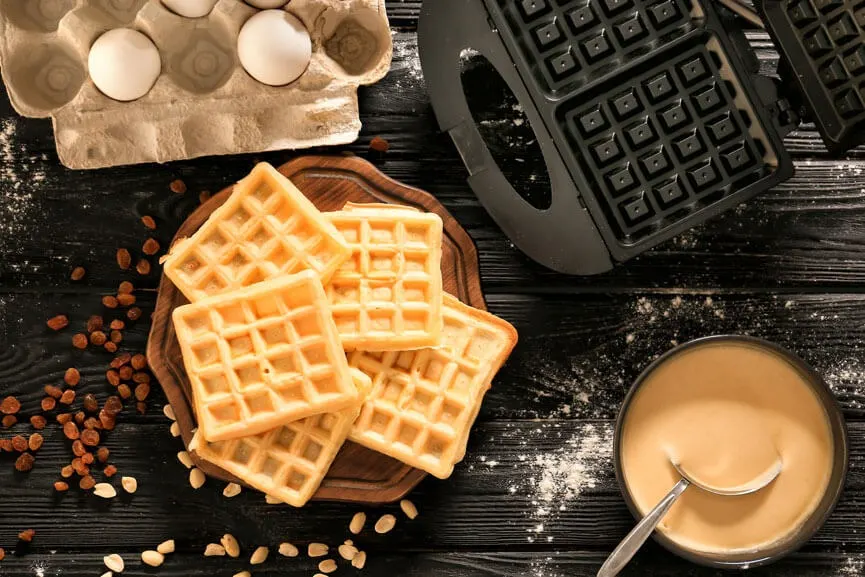 Wafels op een houten plank naast een eierdoos, wafelijzer, kom beslag en zilveren lepel op een donkere houten tafel.