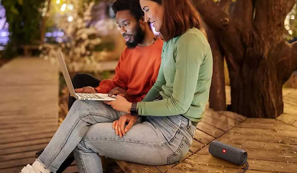 Deux personnes sont assises dehors sur une terrasse en bois avec un ordinateur portable, et une enceinte Bluetooth JBL bleue à côté d’eux.