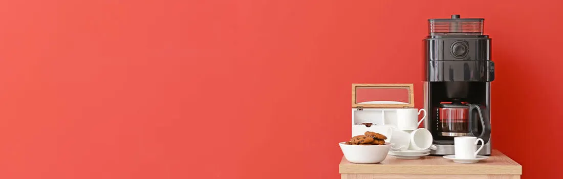 Koffiezetapparaat, kopjes, koekjes en theedoos op een houten tafel tegen een oranje achtergrond.