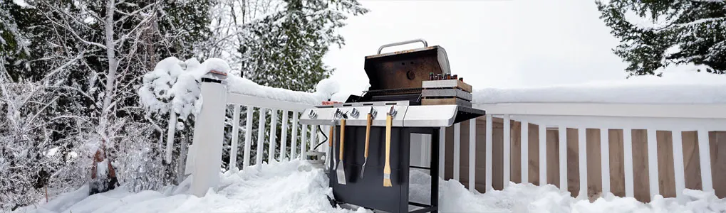 Un barbecue noir est sur une terrasse enneigée, avec une clôture blanche et des arbres enneigés en arrière-plan.