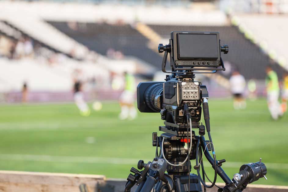 Een zwarte videocamera op een statief filmt een groen voetbalveld met witte lijnen en spelers, op de achtergrond tribunes.