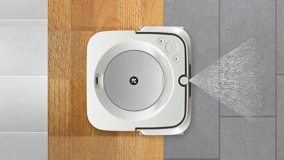 Een witte robot dweilt een grijze tegelvloer, met een houten vloergedeelte aan de zijkant, terwijl er water uit de zijkant spuit.