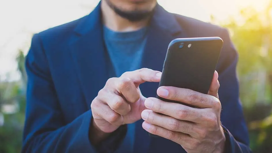Un homme en veste bleue utilise un smartphone noir à l'extérieur, avec un arrière-plan flou de verdure et de lumière du soleil.