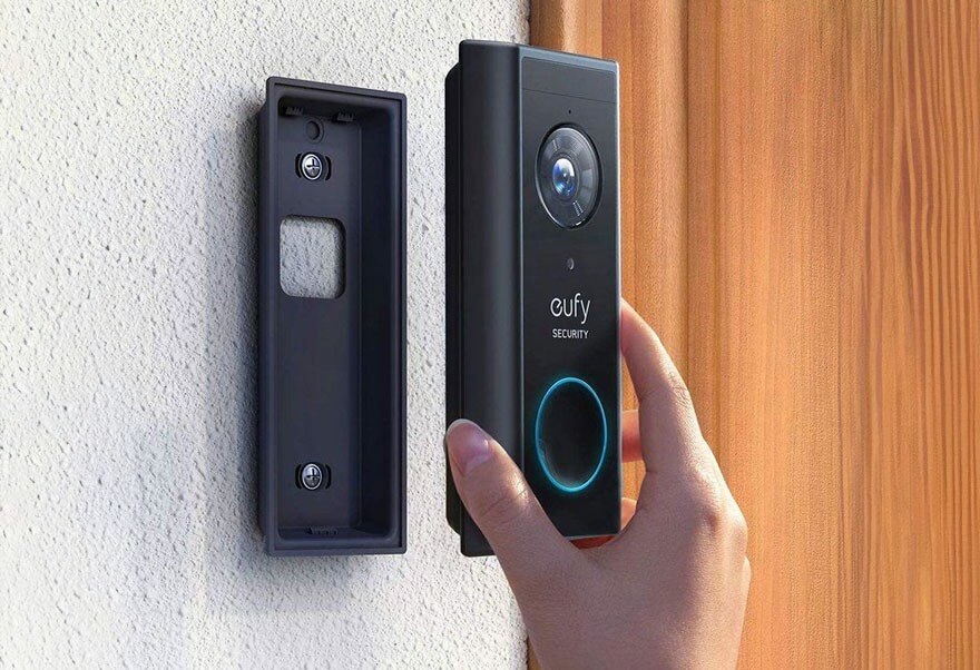 Un boîtier de sonnette noir est fixé au mur blanc. Une sonnette noire Eufy est tenue à la main, près d'une porte en bois.