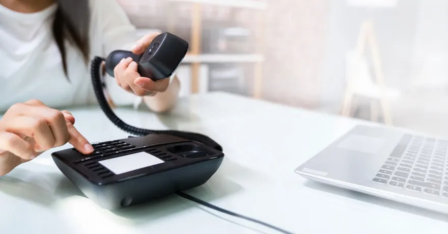 Sur une table blanche, un téléphone noir est utilisé avec une main appuyant sur les boutons. Un ordinateur portable est visible.