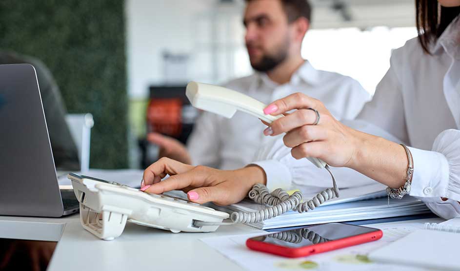 Une personne utilise un téléphone fixe blanc au bureau, avec laptop, papiers et smartphone rouge sur la table, collègues en arrière-plan.