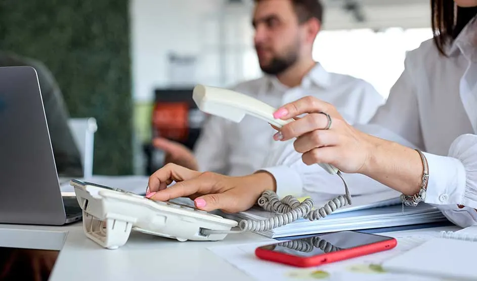 Une personne utilise un téléphone fixe blanc au bureau, avec laptop, papiers et smartphone rouge sur la table, collègues en arrière-plan.