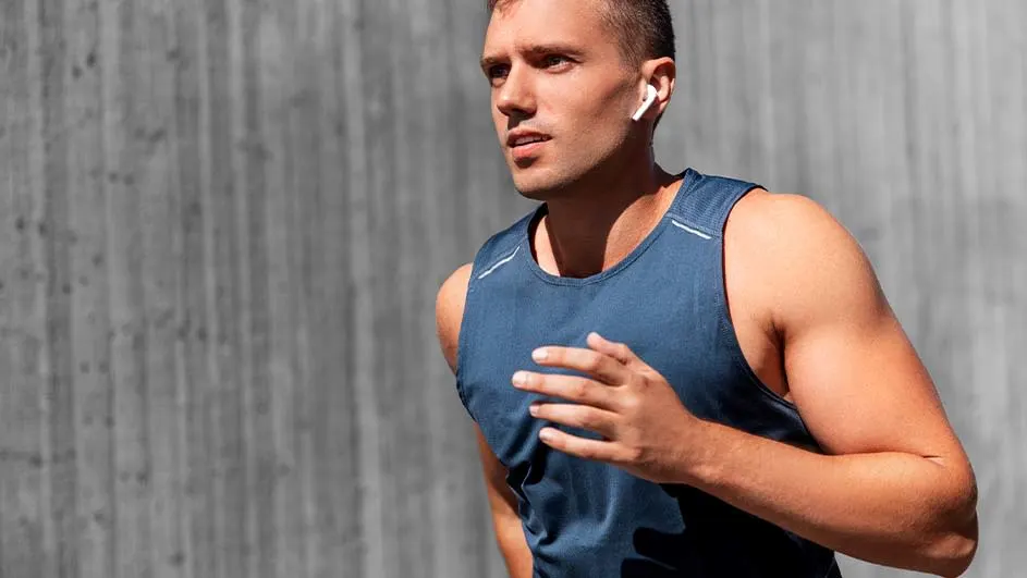 Un homme en débardeur bleu court devant un mur gris. Il porte des écouteurs sans fil blancs.