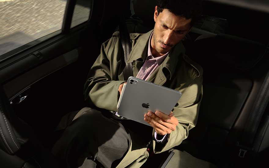 Un homme est assis à l’arrière d’une voiture et travaille avec concentration sur un iPad gris, éclairé par le soleil.