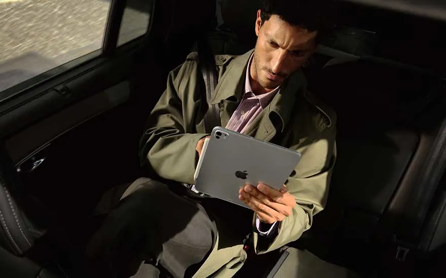 Un homme est assis à l’arrière d’une voiture et travaille avec concentration sur un iPad gris, éclairé par le soleil.