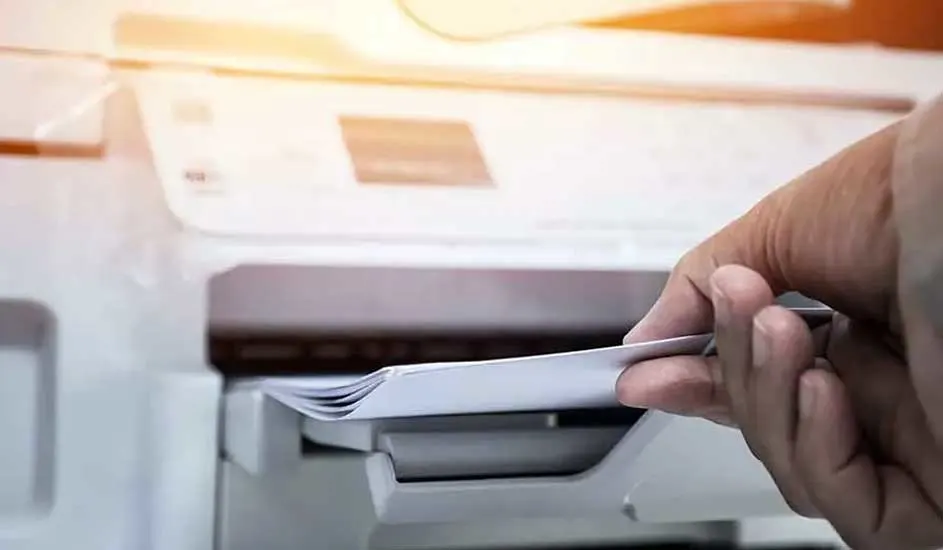 Een hand legt een stapel wit papier in een witte printer. De printer staat op een licht oppervlak.