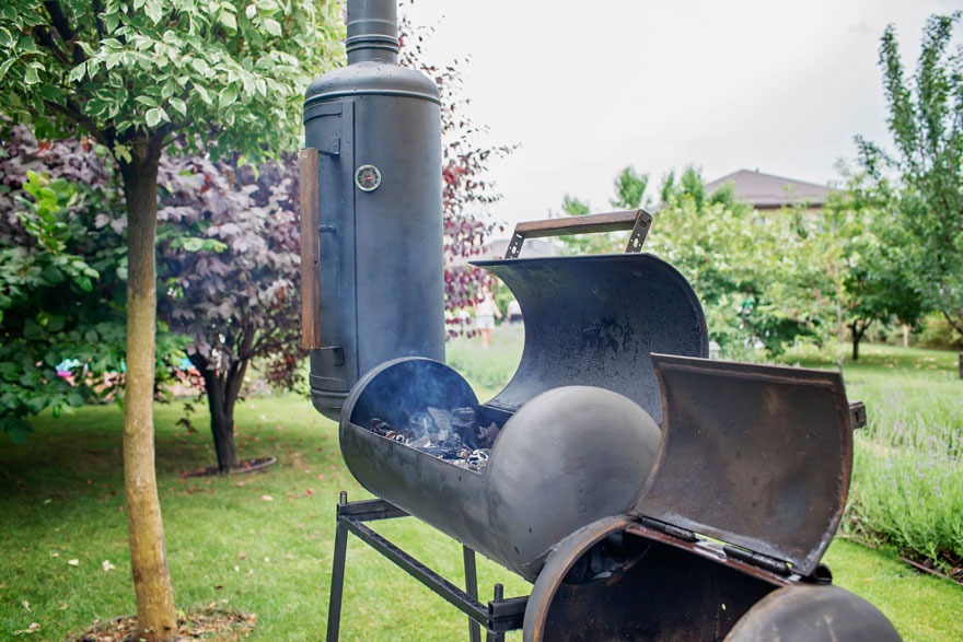 Een zwarte barbecue staat op een gazon met bomen. Rook komt uit de open barbecue.