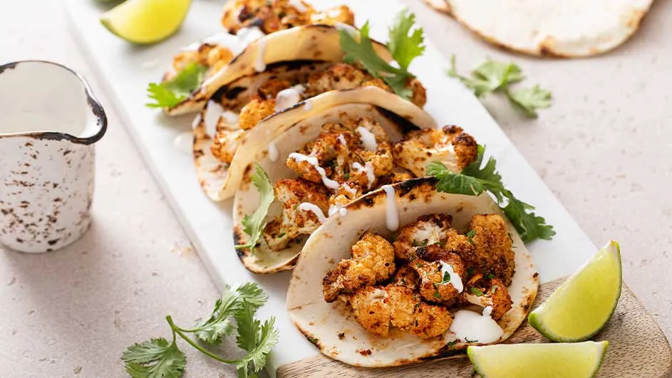 Vier bloemkooltaco's met limoen en koriander op een witte plank. Er staat een witte kan met vlekken naast.