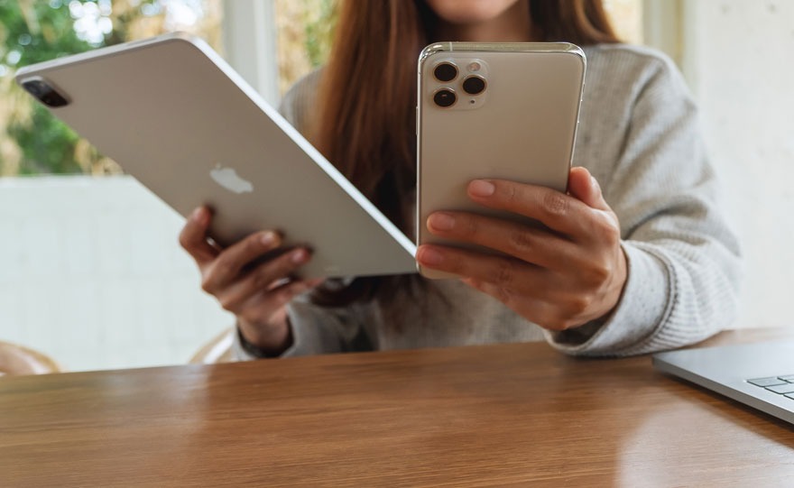Une tablette grise et un smartphone argenté sont tenus au-dessus d'une table en bois, près d'un ordinateur portable.