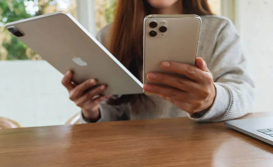 Une tablette grise et un smartphone argenté sont tenus au-dessus d'une table en bois, près d'un ordinateur portable.