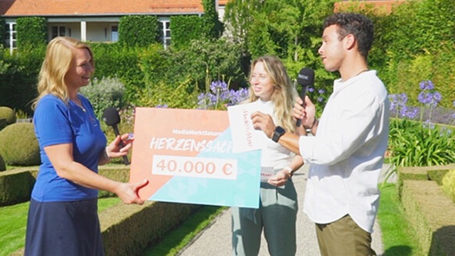 Trois personnes tiennent un chèque de 40 000 € dans un jardin verdoyant avec des fleurs et une maison en arrière-plan.