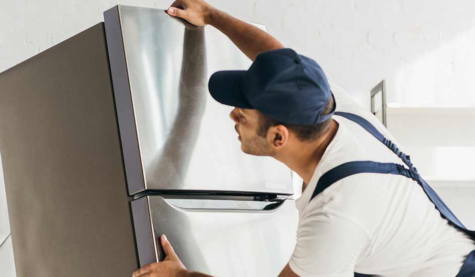 Een man installeert een grote, zilverkleurige koelkast in een ruimte met een witte muur. De koelkast heeft twee deuren.