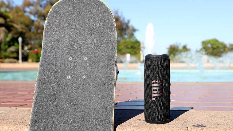 Een skateboard en een zwarte JBL-luidspreker staan buiten, voor een fontein en een blauw zwembad.