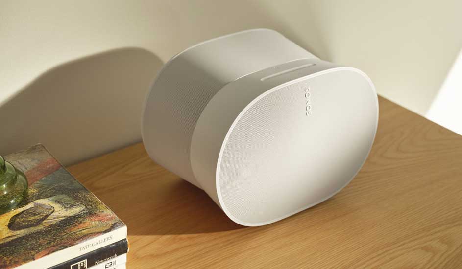 Haut-parleur blanc sur table en bois, livres et ombre au mur. Lumière douce.