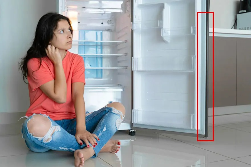 Une cuisine avec un frigo blanc vide, porte ouverte. Une personne assise sur le sol carrelé en avant-plan.
