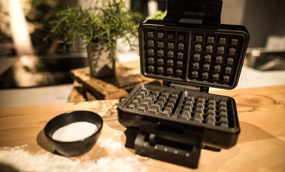 Une gaufrier noir ouvert sur une table en bois, avec un bol noir rempli de sucre et une plante verte en arrière-plan.