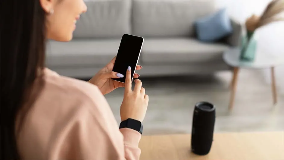 Une personne utilise un smartphone près d'un haut-parleur noir sur une table en bois. Un canapé gris est visible en arrière-plan.