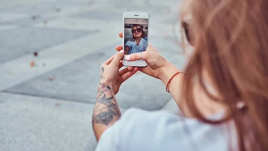 Een vrouw met lang haar houdt een witte telefoon vast. Op het scherm is een foto te zien. De achtergrond is een grijze stenen ondergrond.