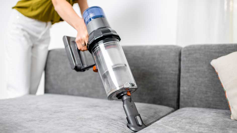 Une personne utilise un aspirateur à main sur un canapé gris clair dans une pièce lumineuse.